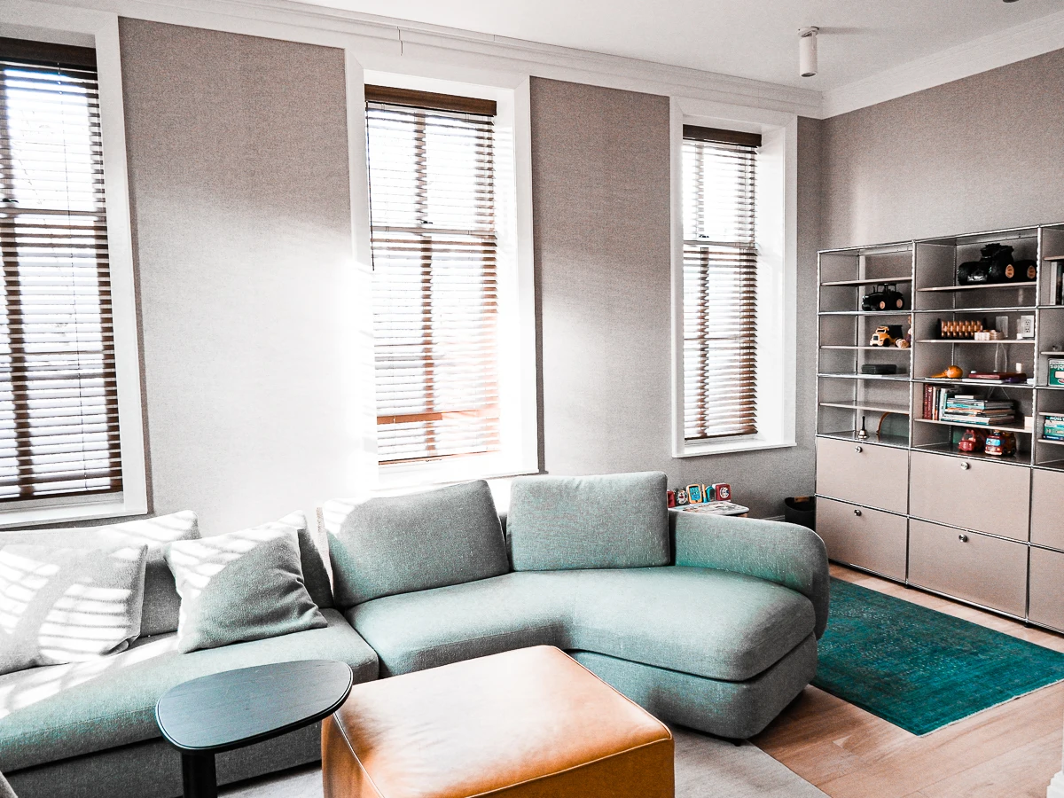 Modern living room renovation by ZS NY Improvement featuring curved gray sectional sofa, cognac leather ottoman, teal rug, modular metal shelving, wooden blinds, and light hardwood floors